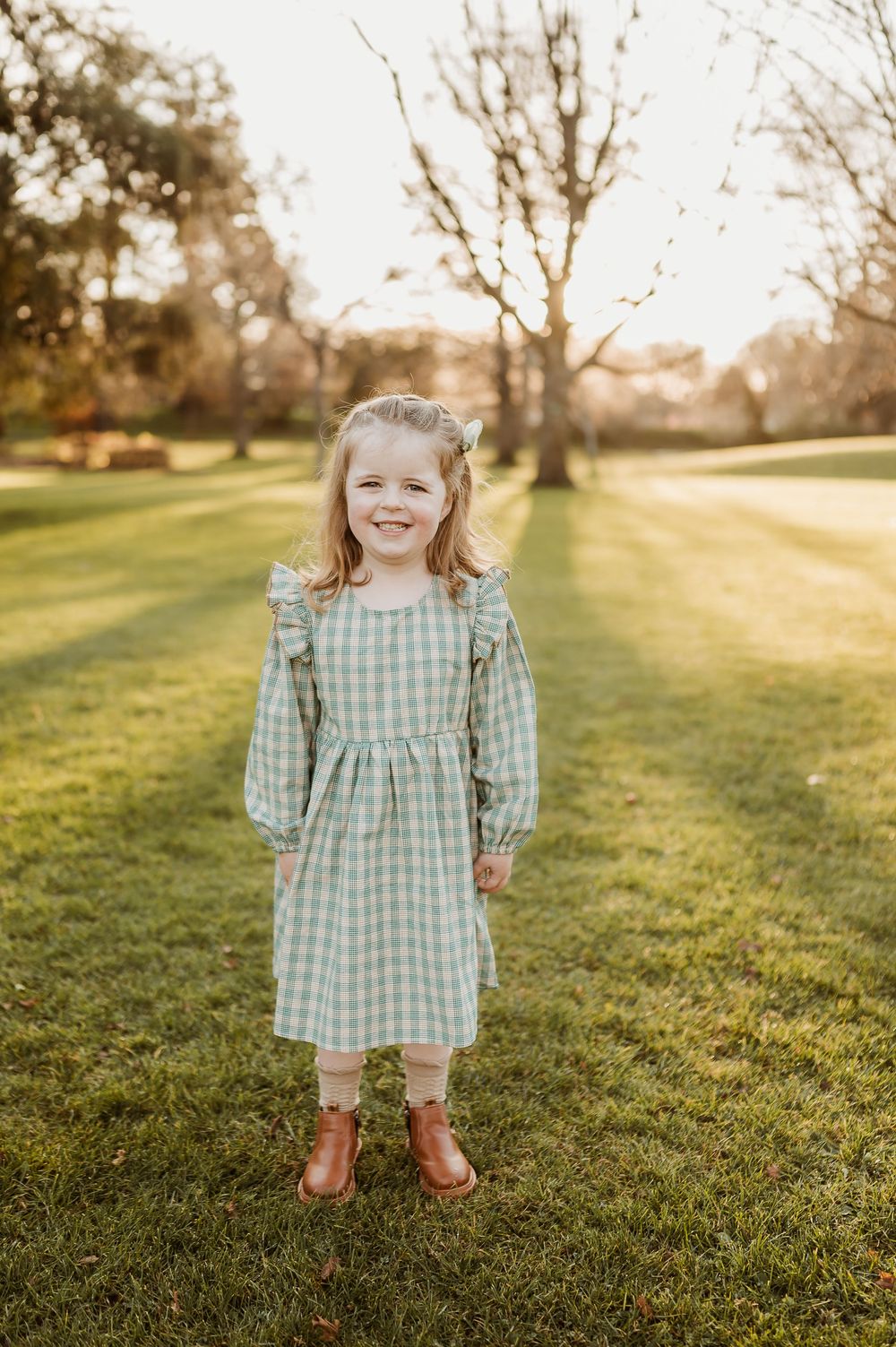 A child stands in a green gingham dress and brown boots on sunlit grass during golden hour with bare trees in background.