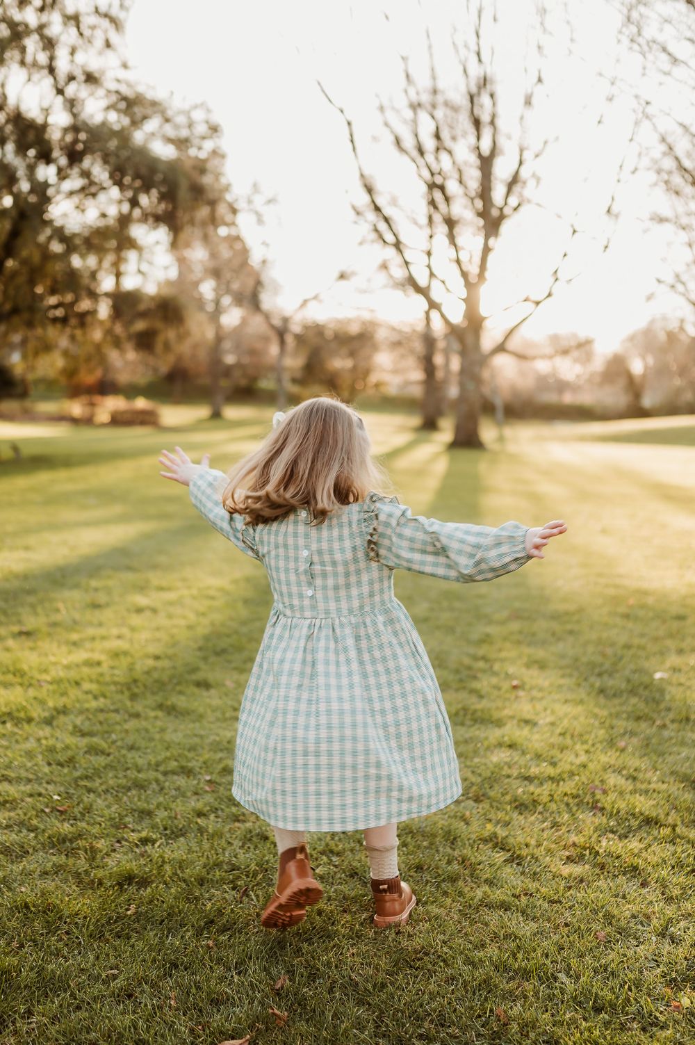 Small child in a checkered dress twirls joyfully on green grass during golden hour sunset.