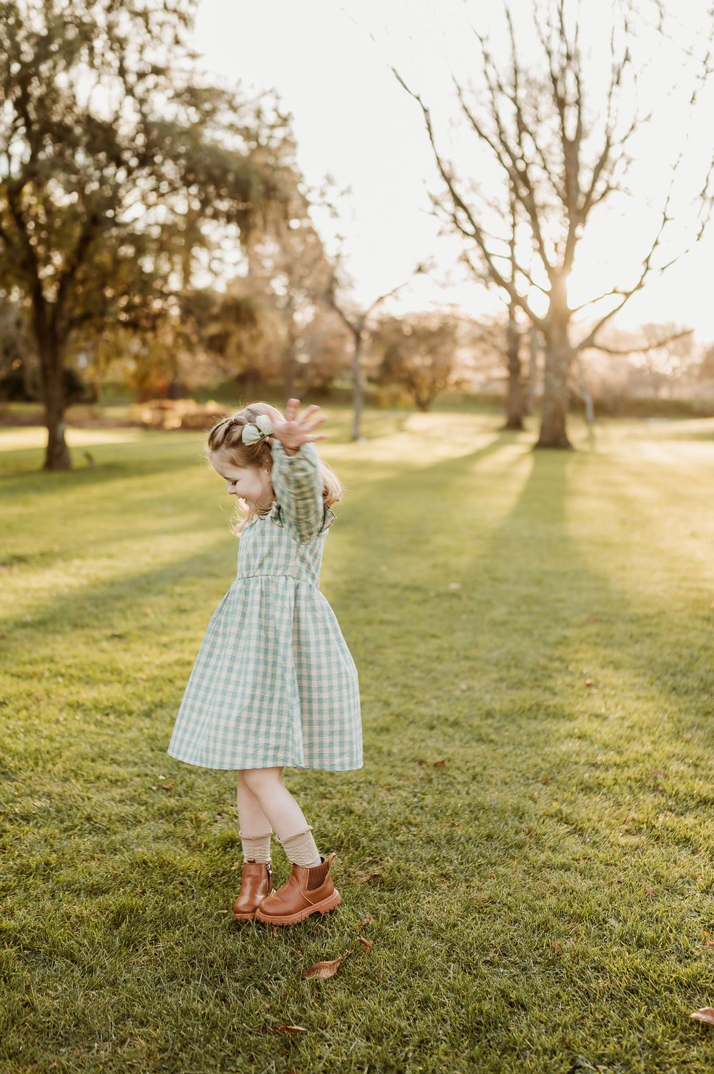 Young child in a gingham dress twirling playfully on grass during golden hour.
