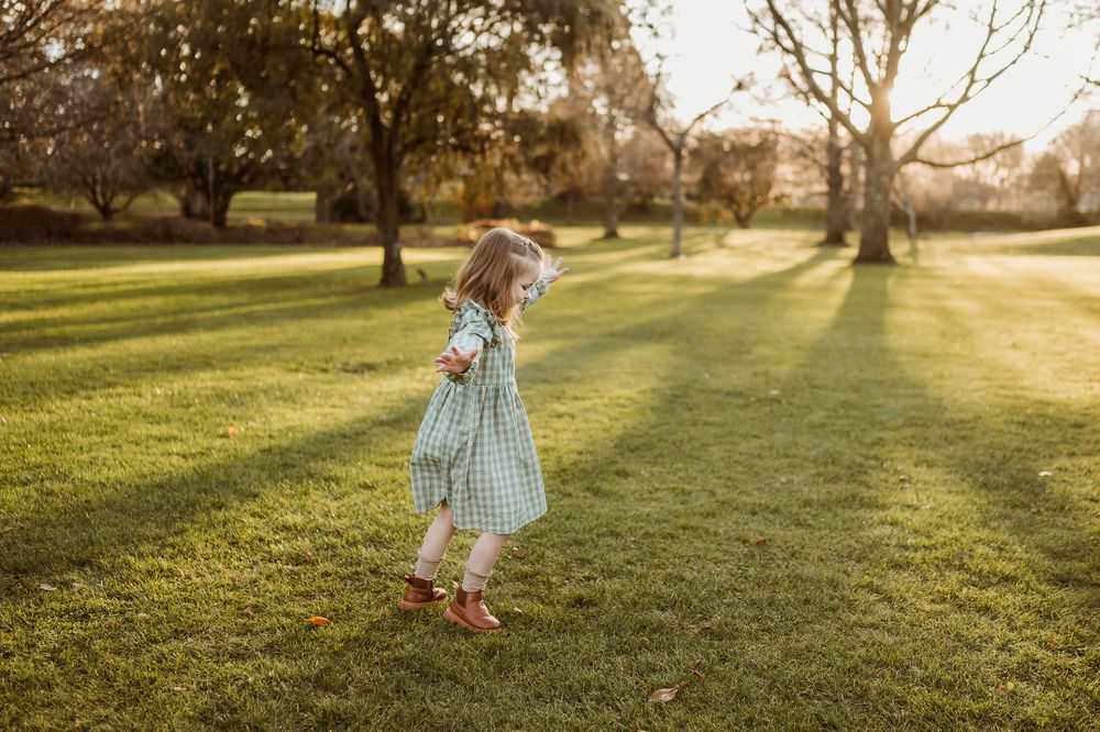 A child in a light blue dress spins playfully on a sunlit grass lawn during golden hour.