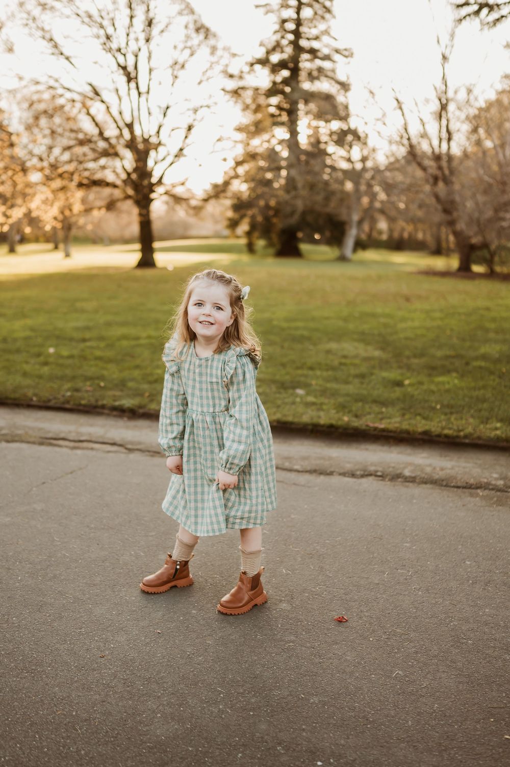 A child in a green gingham dress and brown boots stands on a paved path in a park during golden hour.