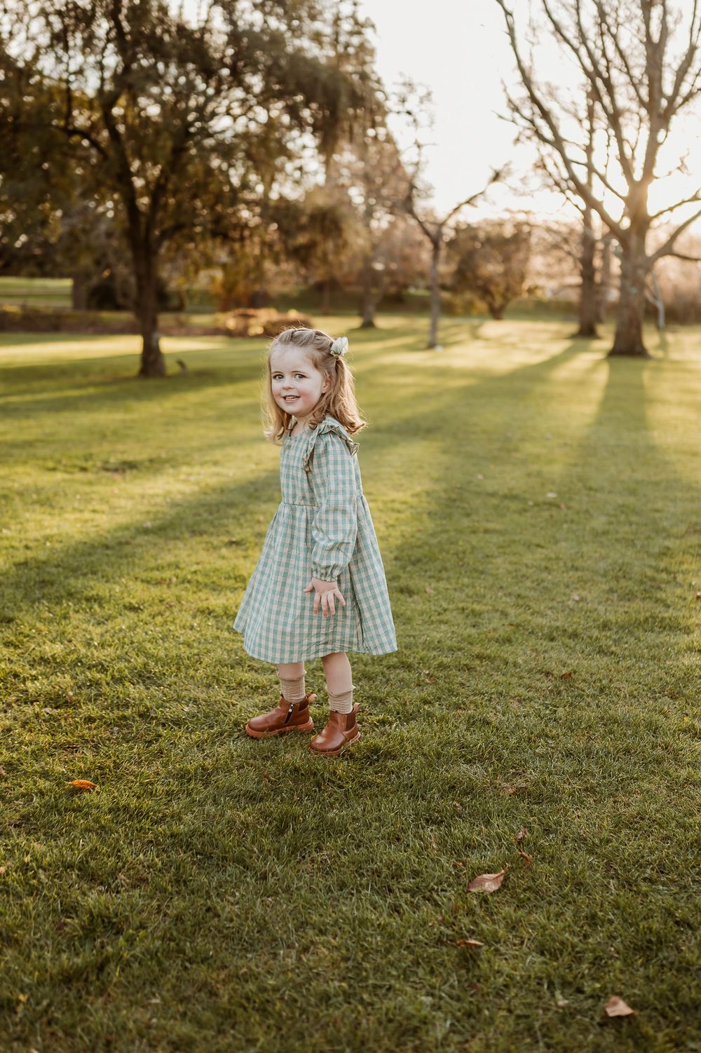 A child wearing a checkered dress smiles while standing on grass at sunset.