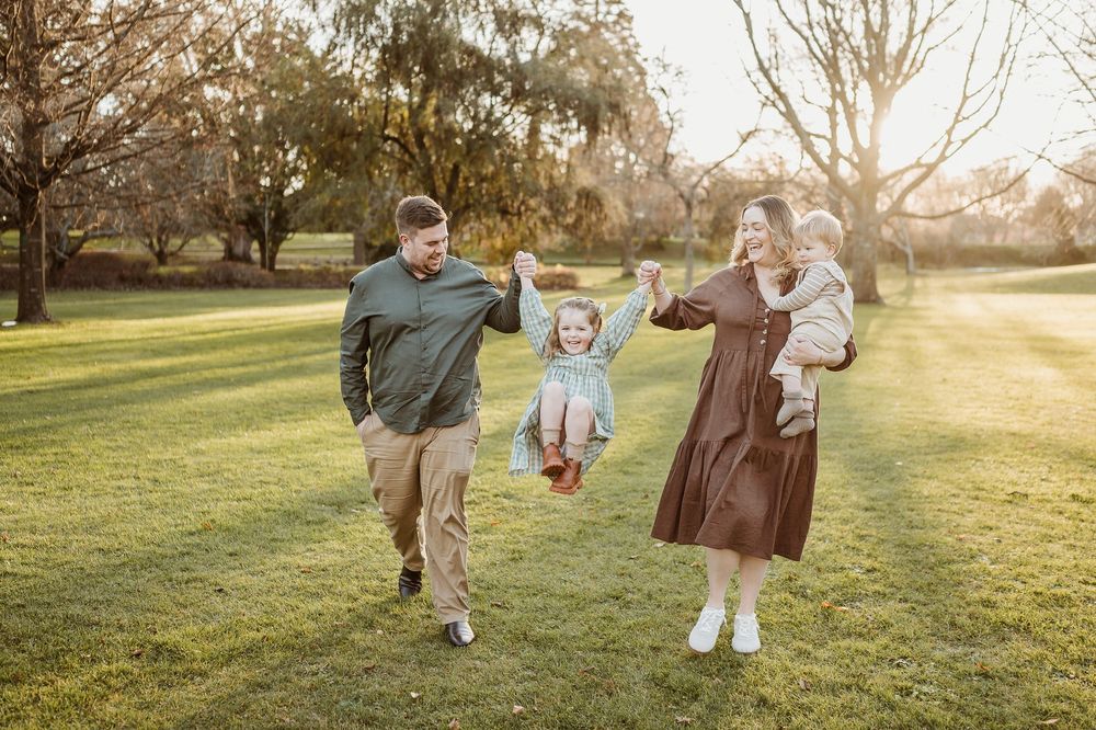 A family walks together in a sunlit park during golden hour, holding hands and enjoying a playful moment on the grass.