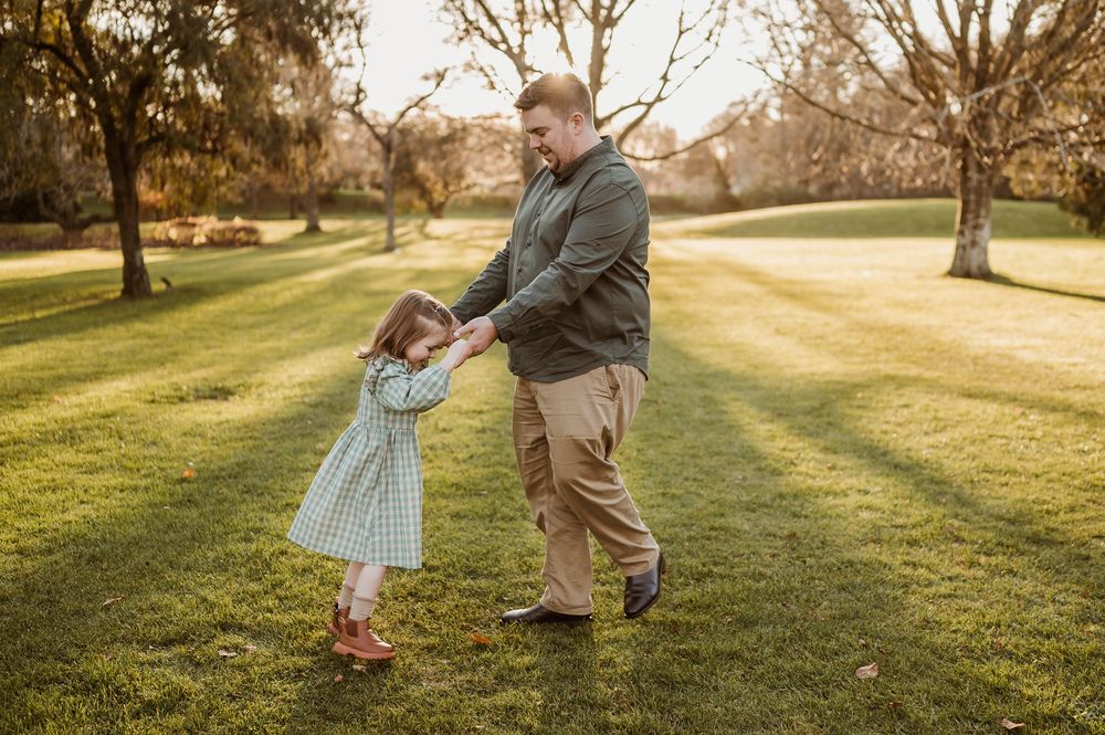 A playful moment between adult and child dancing together on a sunlit lawn in a park during golden hour.