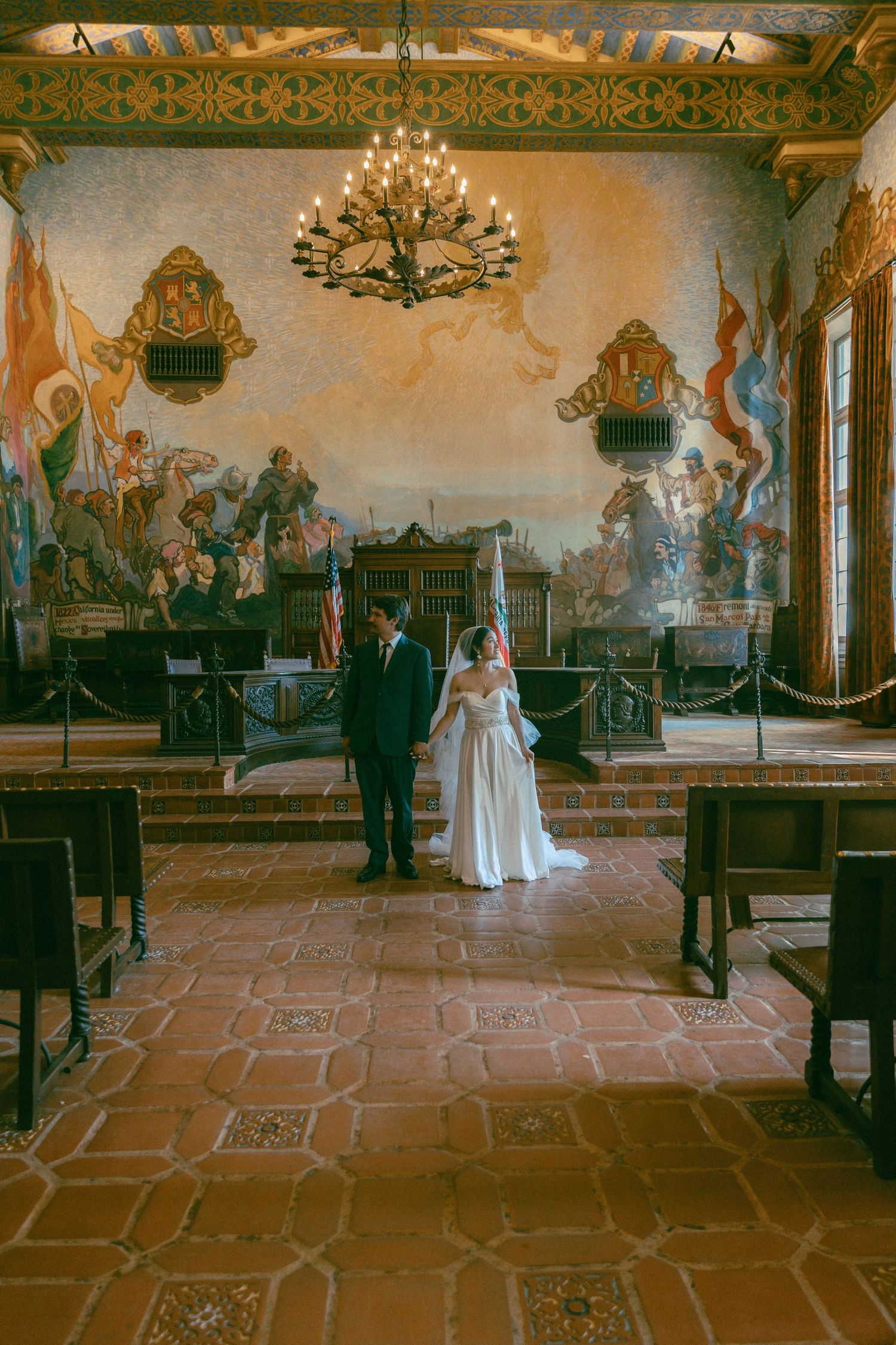 bride and groom stand in the historic mural room with ornate murals, a chandelier and tiled floors at Santa Barbara Courthouse.