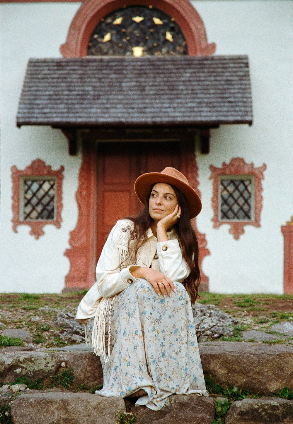 A woman in a floral dress sits on stone steps before an old wooden door, gazing softly into the distance.
