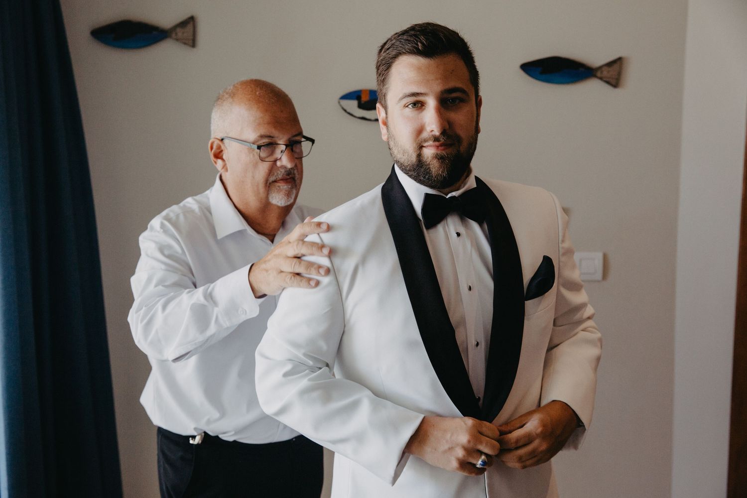 A groomsman helps adjust the groom's bowtie while preparing for a formal wedding event.