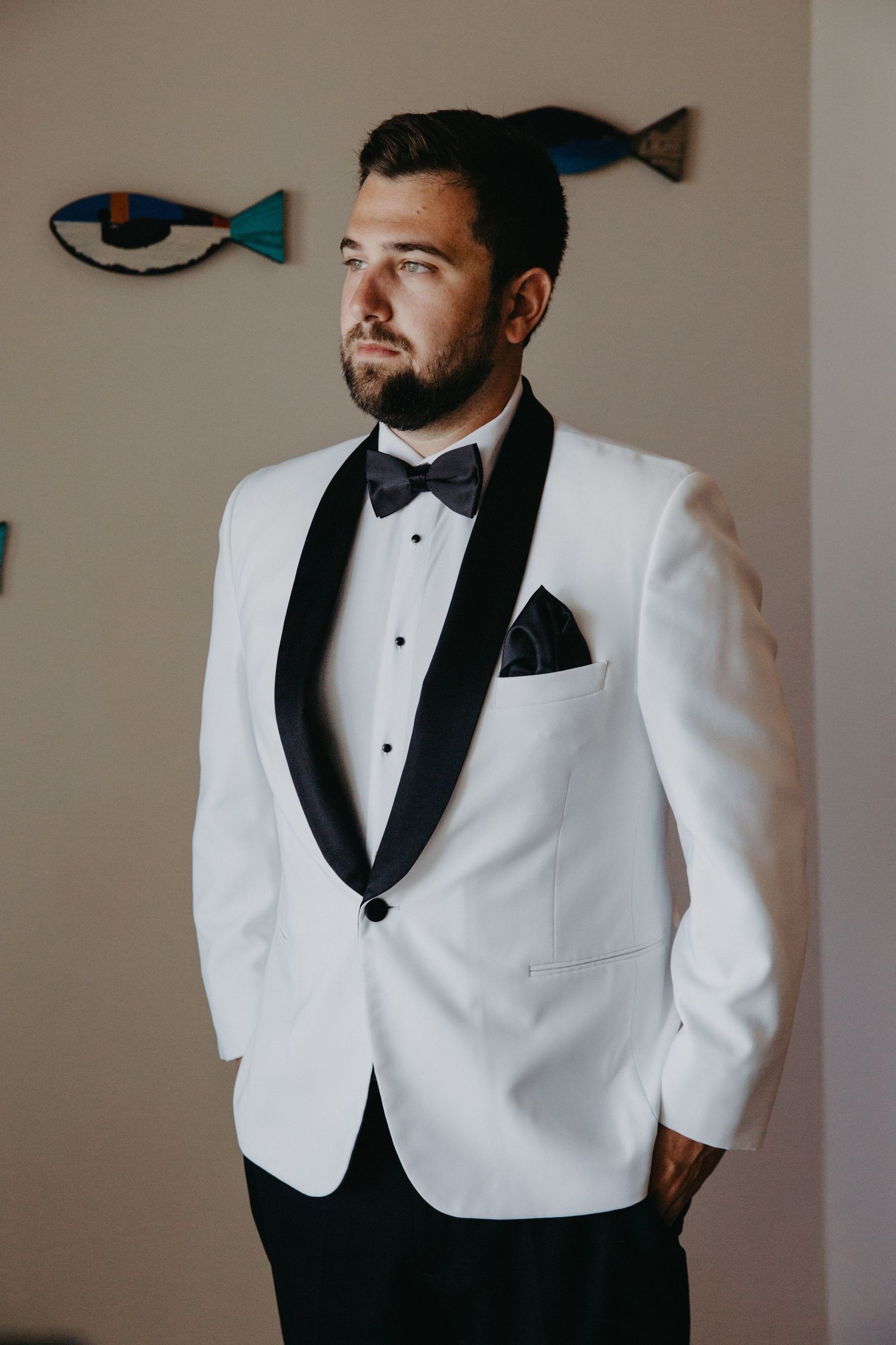 A classic white dinner jacket with black shawl lapel and bow tie stands elegantly against a neutral wall with fish decor.