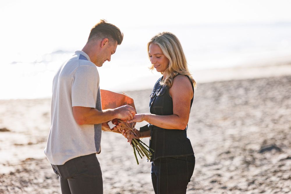 A romantic proposal scene unfolds on a sunlit beach with ocean waves in the background.