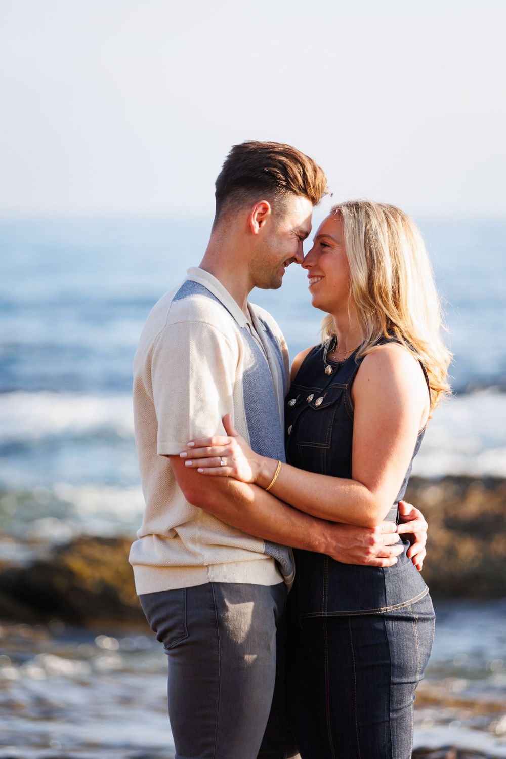 A couple shares a tender moment while embracing on a rocky beach with ocean waves in the background.