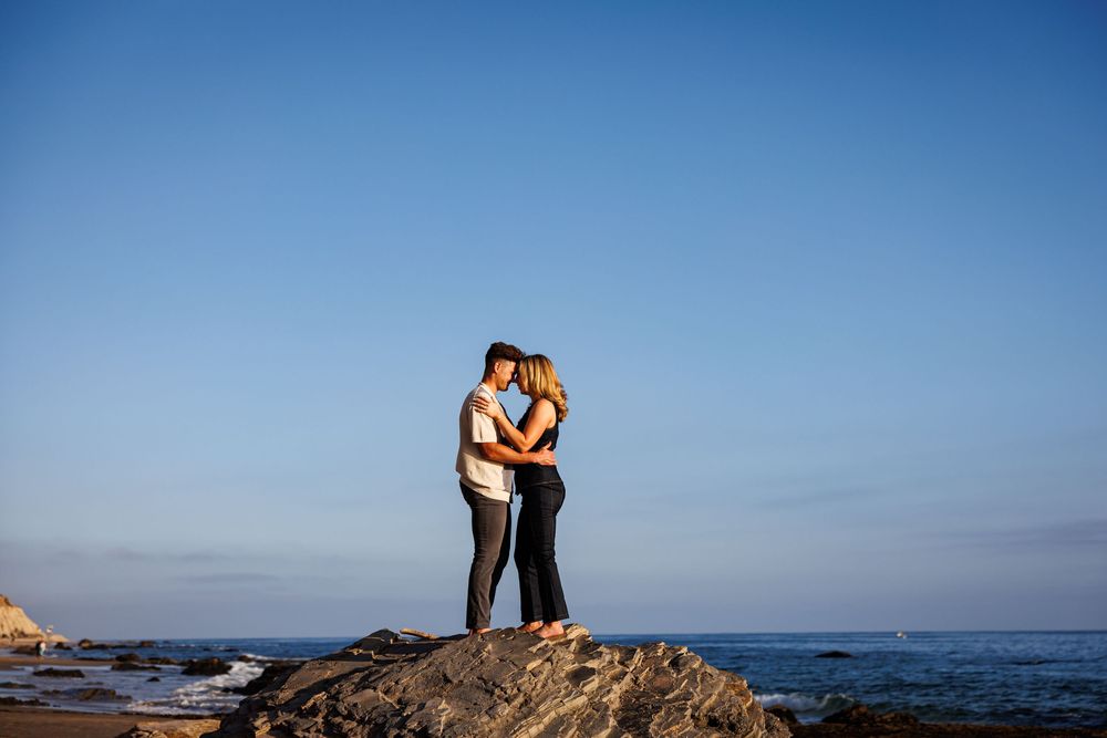 Couple embraces at sunset on rocky coastal cliff overlooking blue ocean in Crystal Cove.