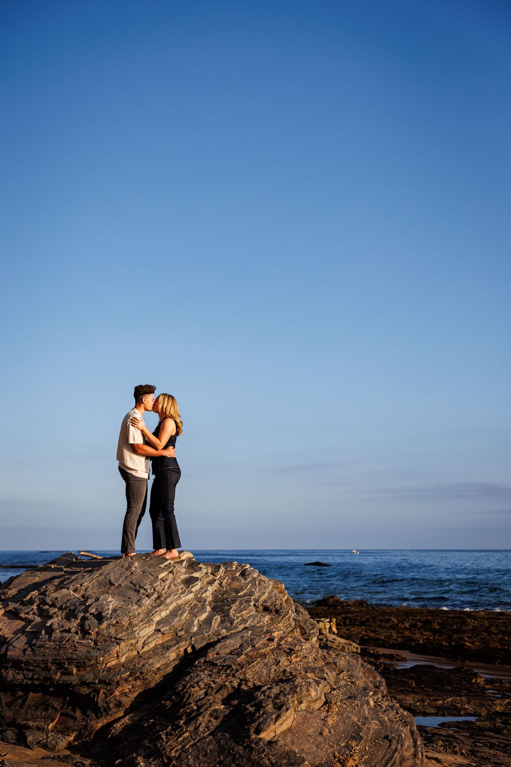 A couple embraces while standing on a rocky ocean outcrop against a vibrant blue sky at sunset.