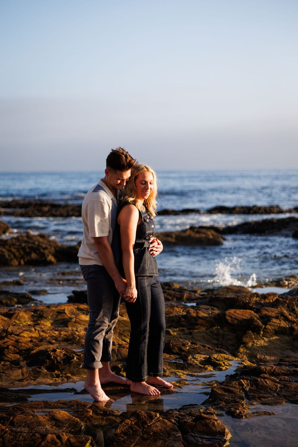 A couple shares intimate moments along a rocky beach shoreline at sunset, creating a romantic coastal scene.