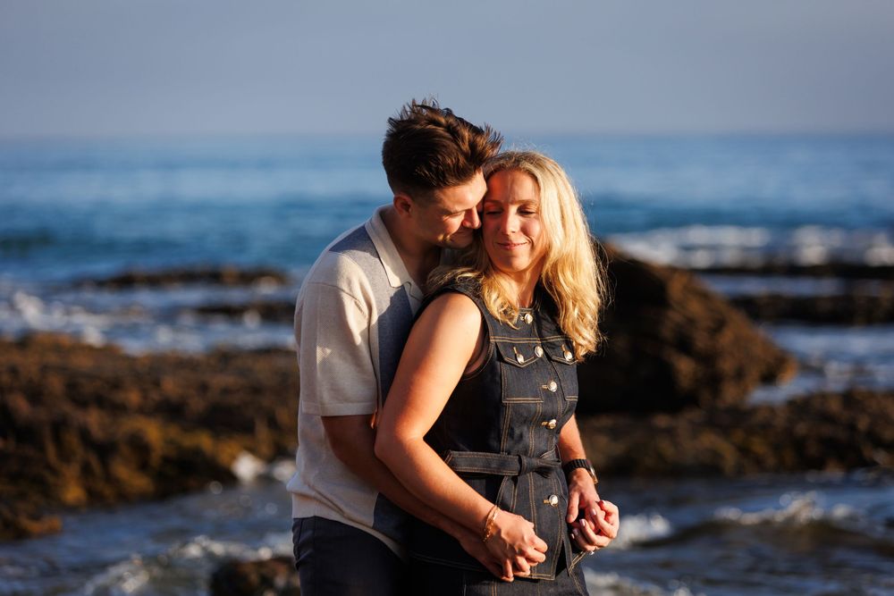 A couple shares a romantic moment on rocky beach shores at sunset overlooking the ocean waves.