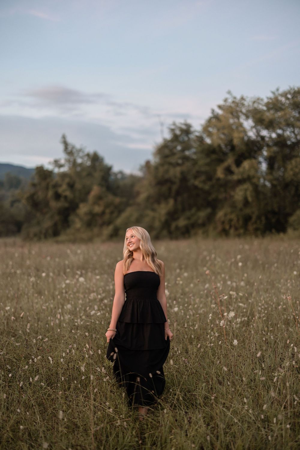 A person in a black dress stands in a wildflower meadow at dusk with mountains and trees in the background.