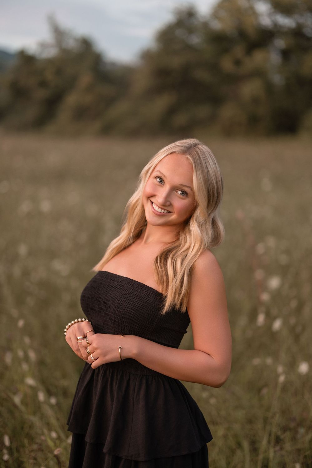 A model in a black dress poses playfully in a golden field during sunset with a natural, candid expression.