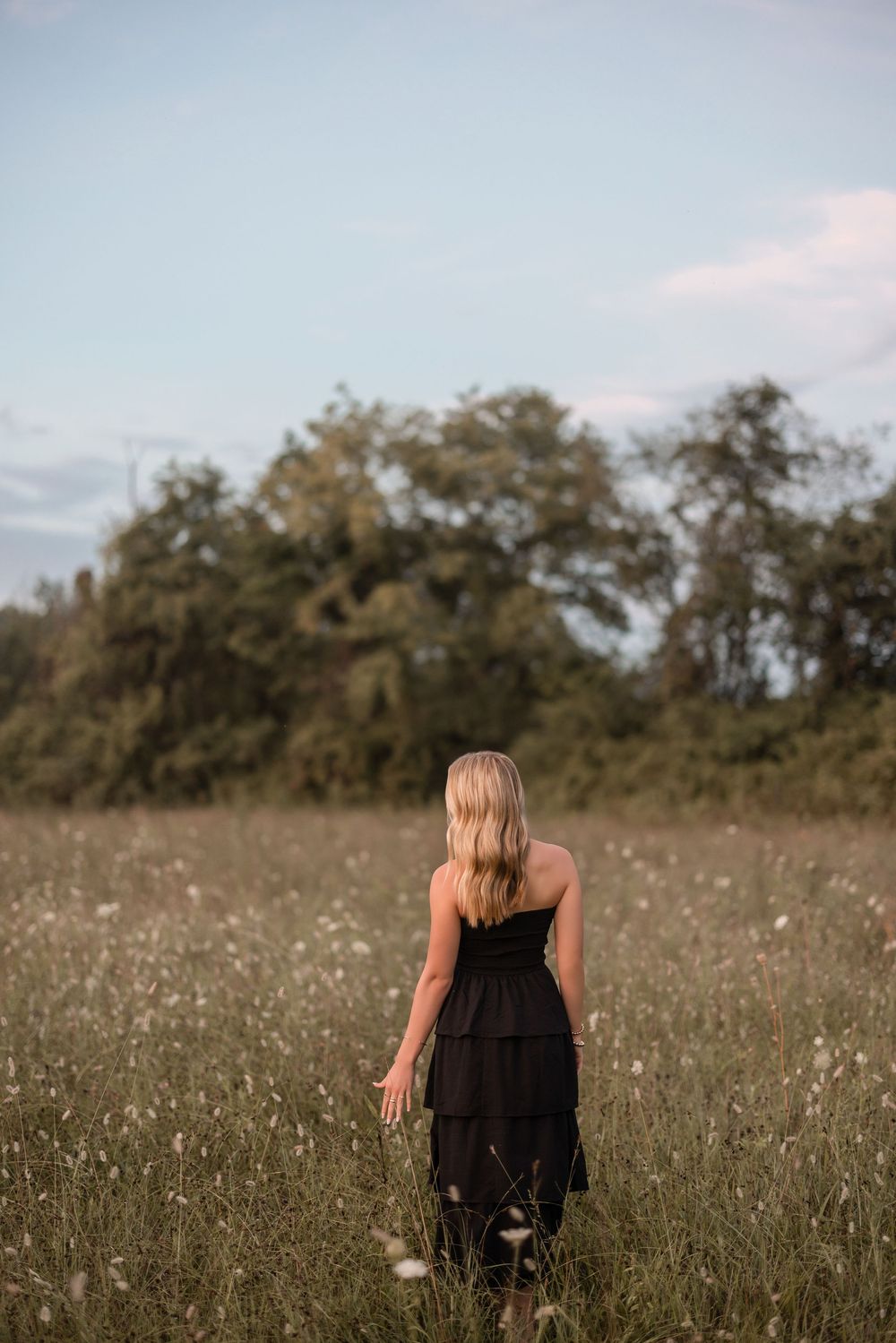 A silhouette figure in a black dress stands in a golden wildflower meadow at sunset with trees in background.