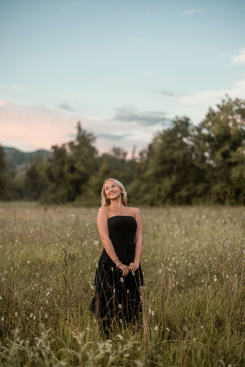 Woman in black dress poses elegantly in a wildflower meadow at sunset with mountains in background.
