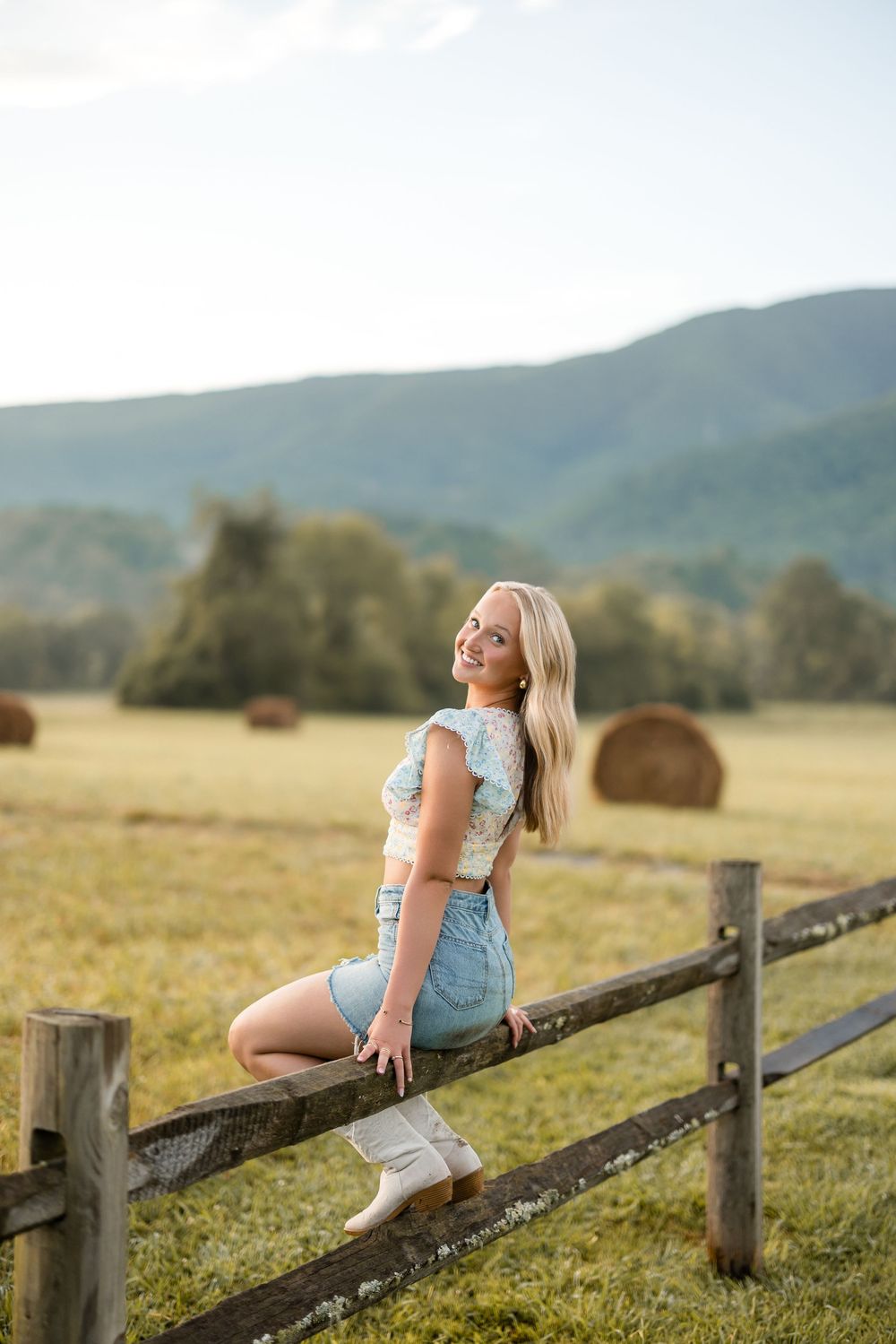 Person in denim overalls sits on wooden fence in countryside with hay bales and mountains in background at sunset.