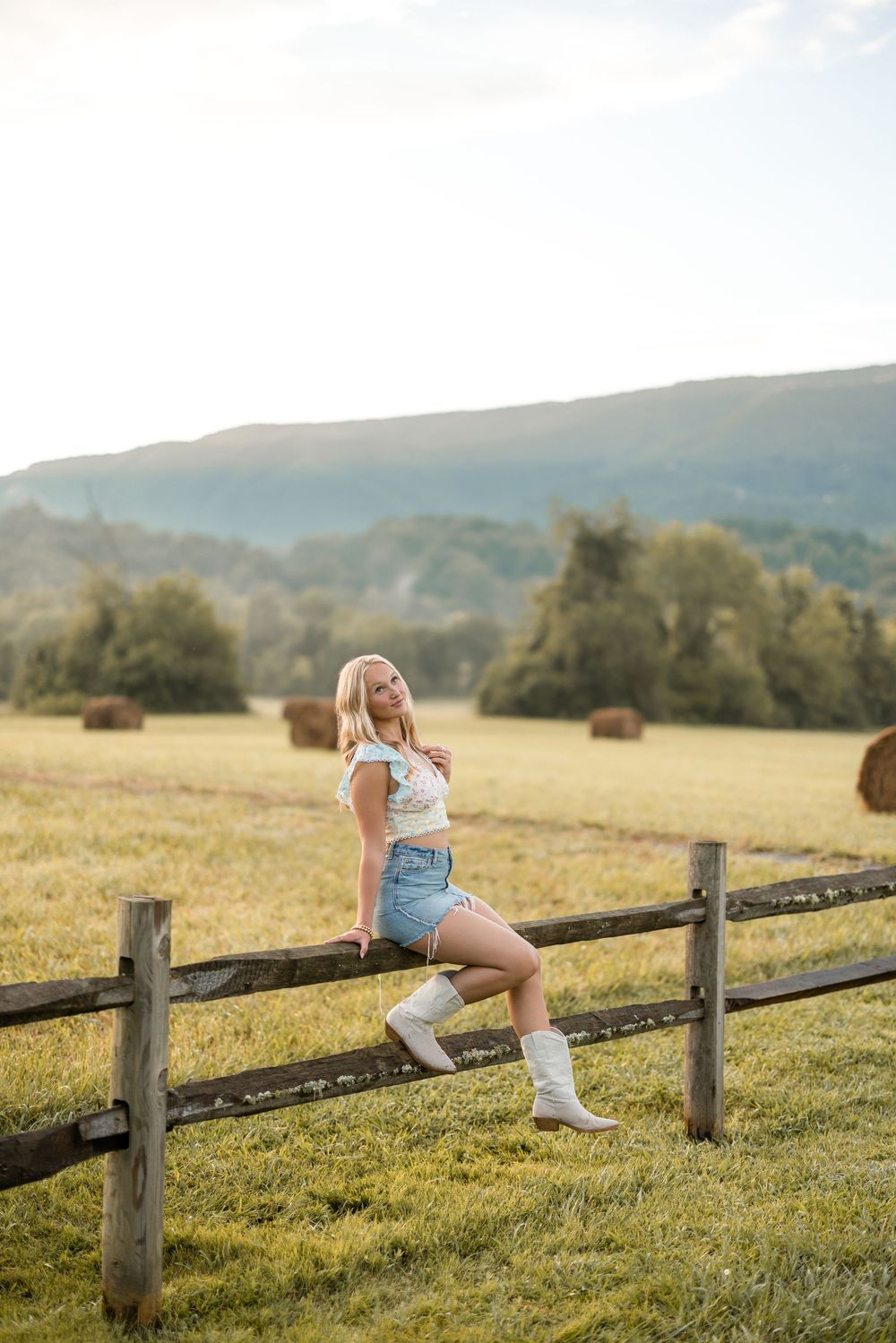 Figure in denim and white socks sits casually on rustic wooden fence with rolling hills in background.