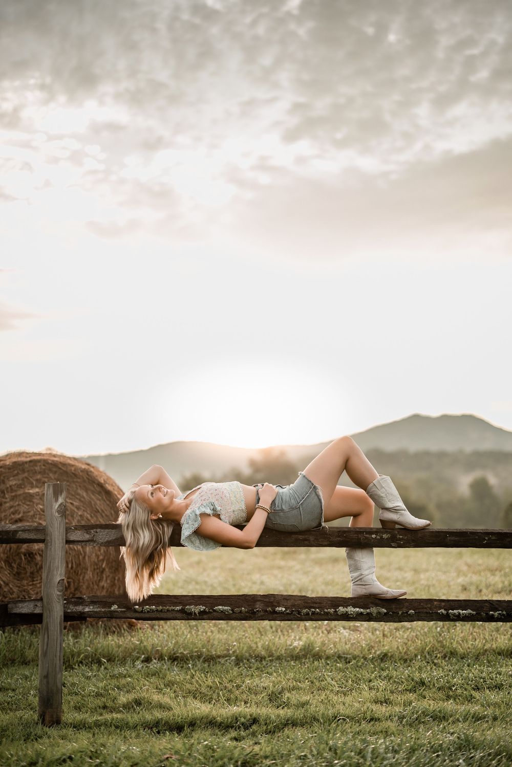 Person relaxes on rustic wooden fence against sunset mountain landscape with hay bales visible in field.
