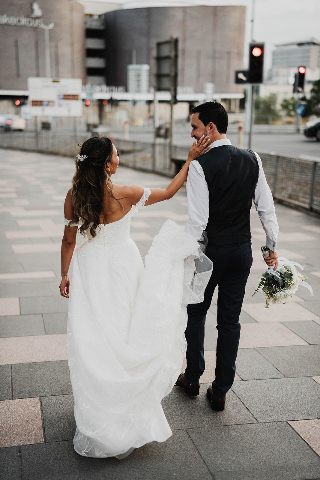A couple in wedding attire dances playfully on a city sidewalk, with the bride's white dress flowing in motion.