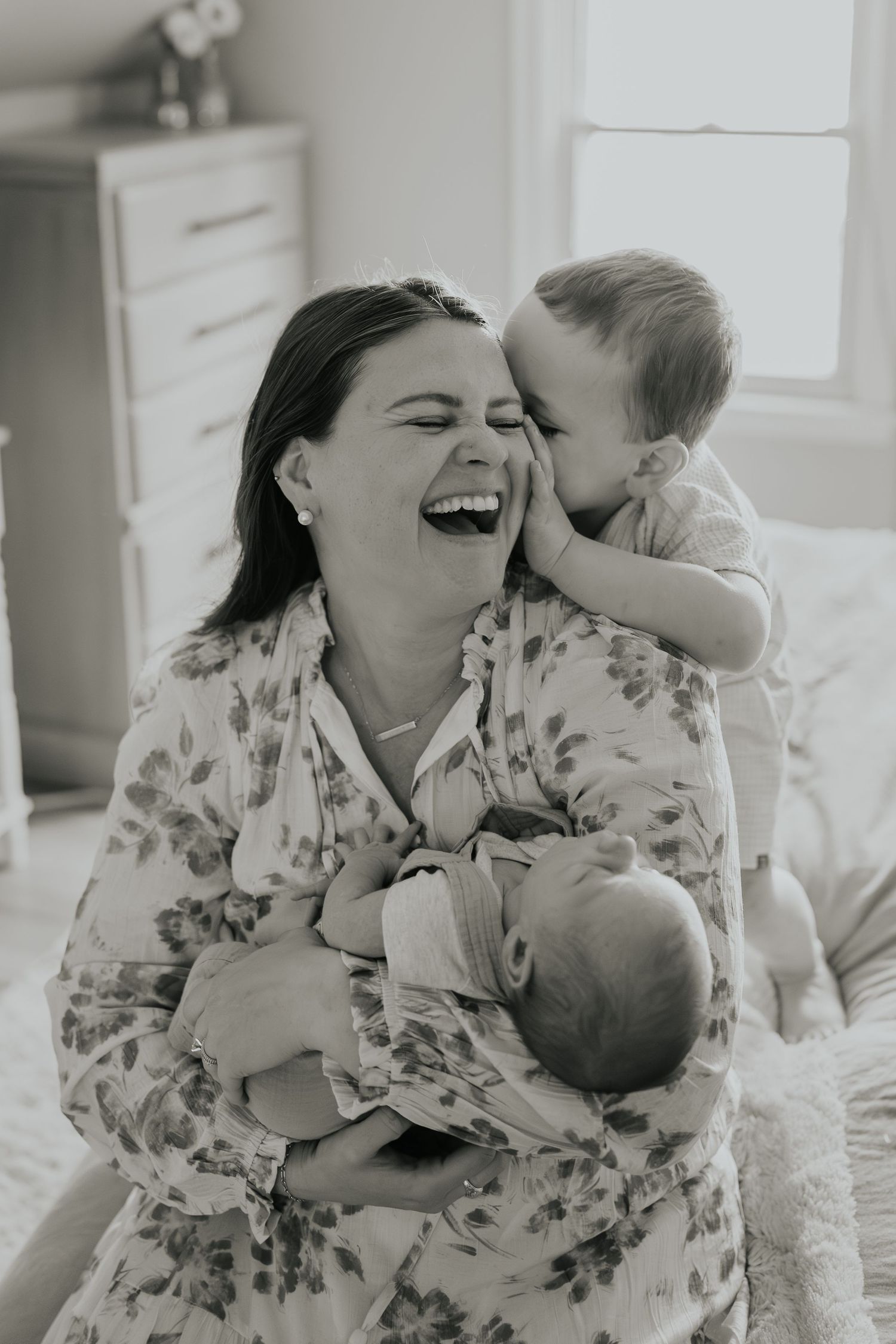 Black and white photo capturing a joyful moment between a parent and children on a bright bedroom bed.