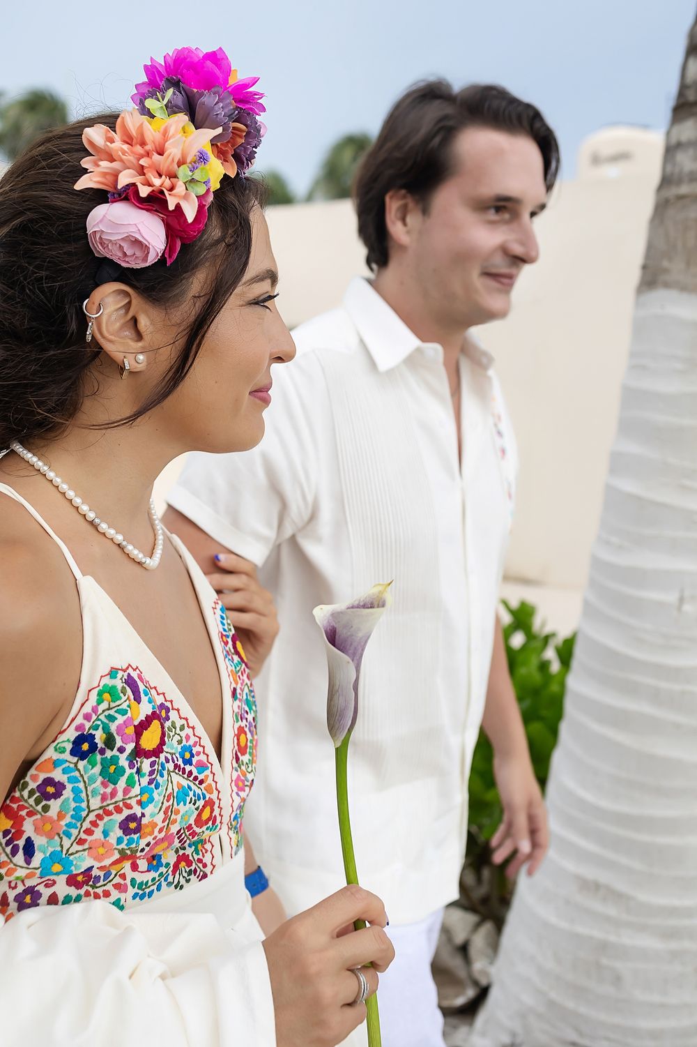 A couple in white attire at a tropical wedding, with the bride wearing vibrant floral hair accessories.