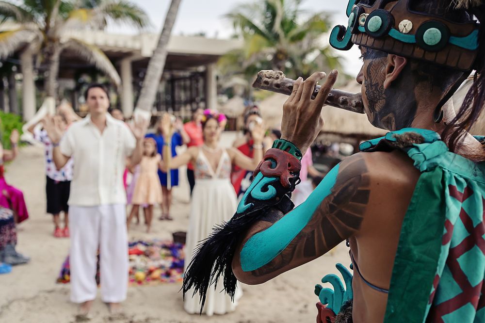Traditional Mayan performer in turquoise costume plays flute at a beachside event with palm trees and tourists.