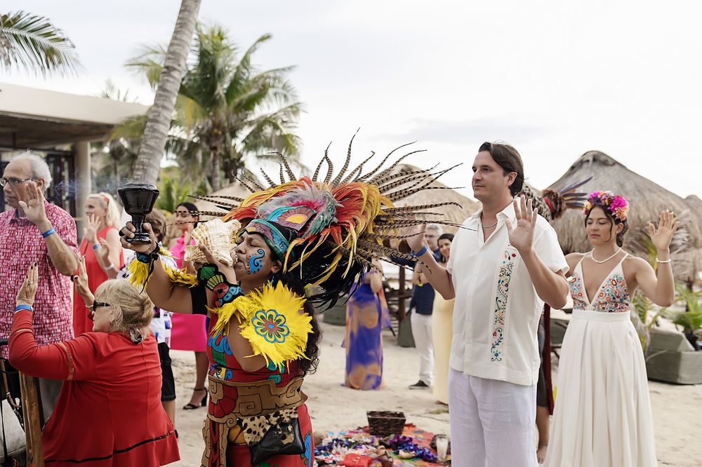 Bride walks down beachfront wedding aisle while guests fan themselves with colorful paper fans in tropical setting.