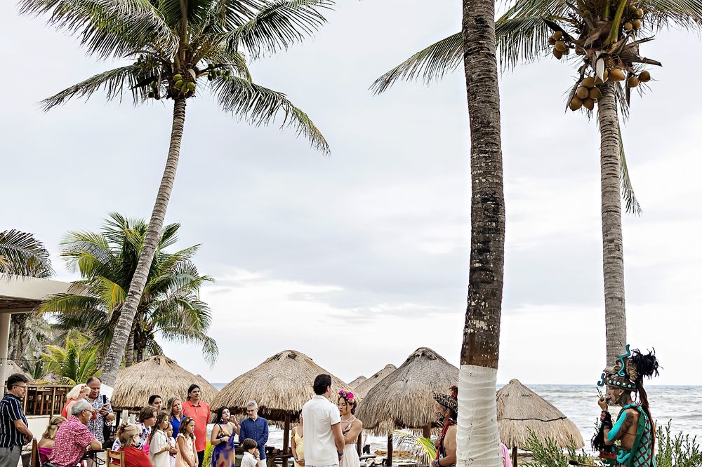 A beachside wedding ceremony under palm trees and thatched-roof huts with guests in attendance.