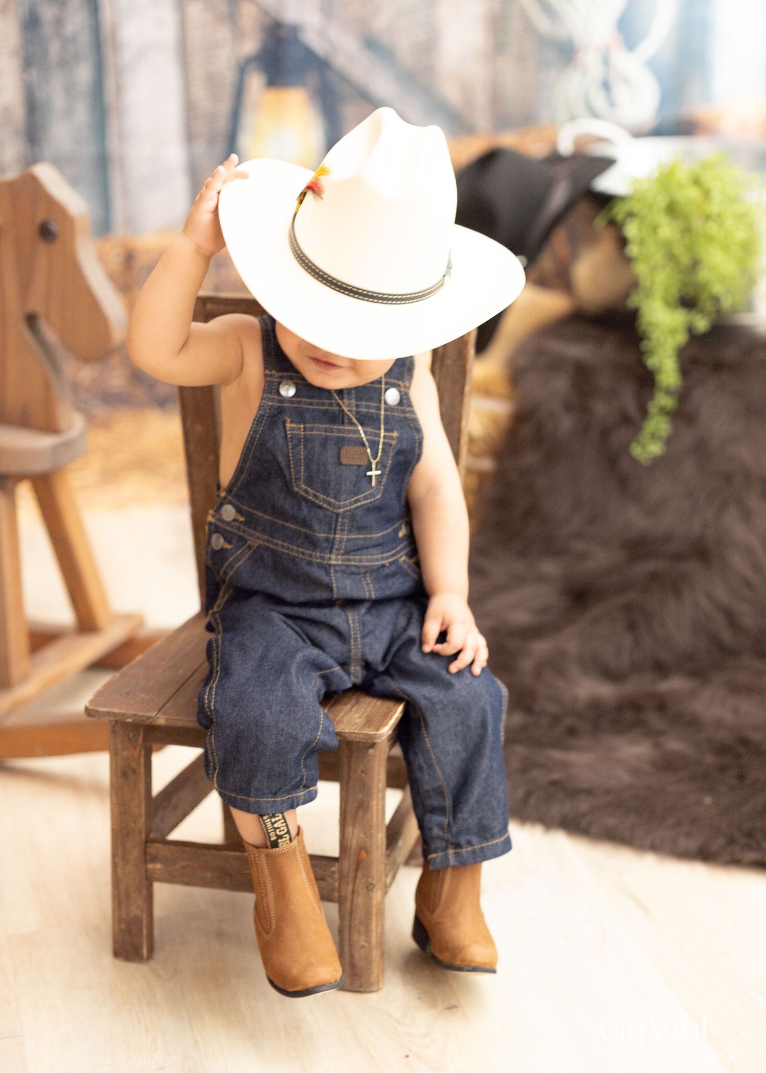 A small child in denim overalls and a white cowboy hat sits on a rustic wooden chair wearing brown boots.