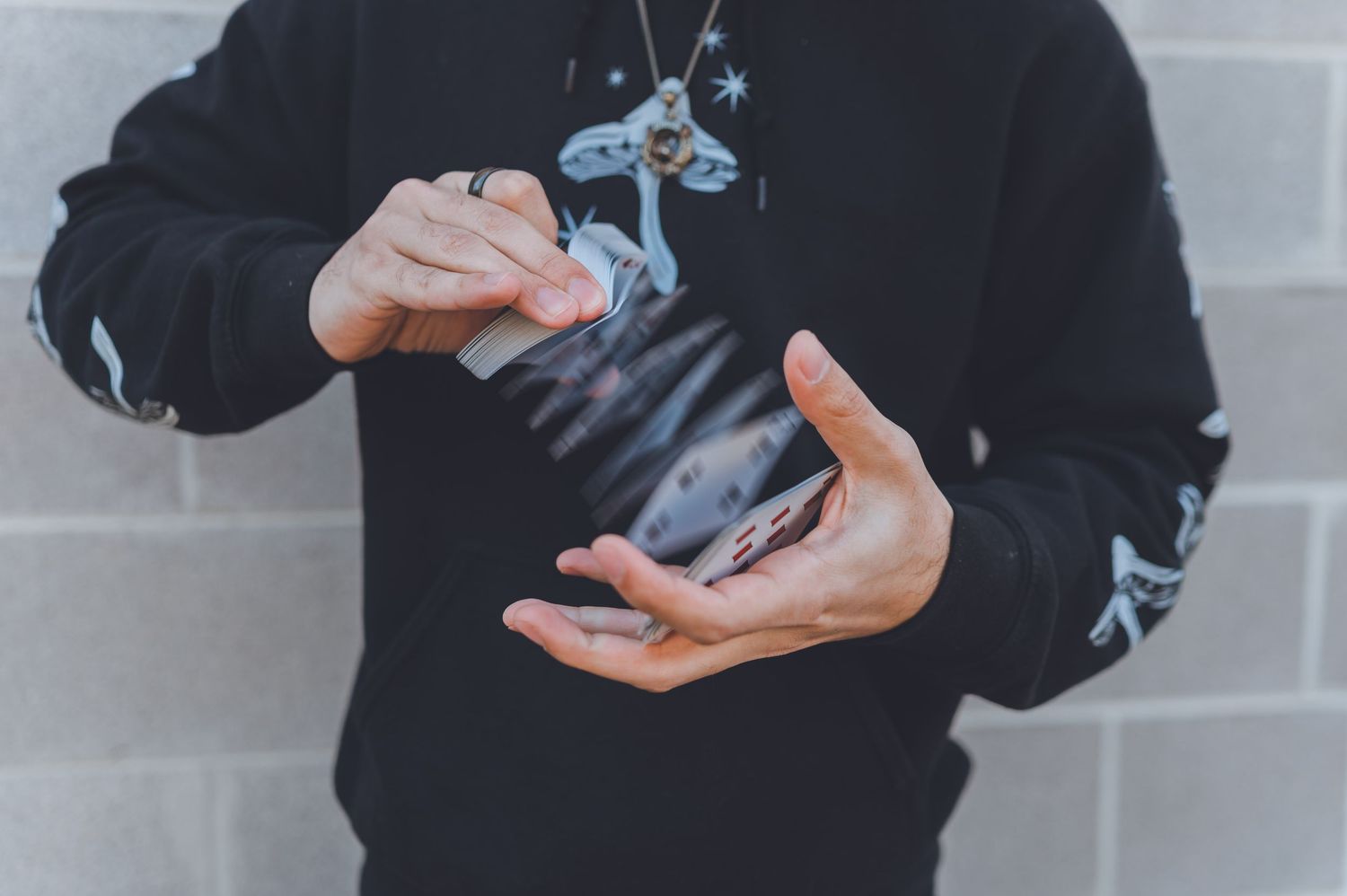 Card magician in black hoodie performs fanning technique with deck against grey wall.