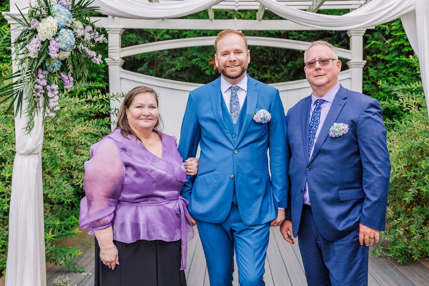 Wedding party poses under a white decorated arch in a garden setting, with blue suits and lilac attire.
