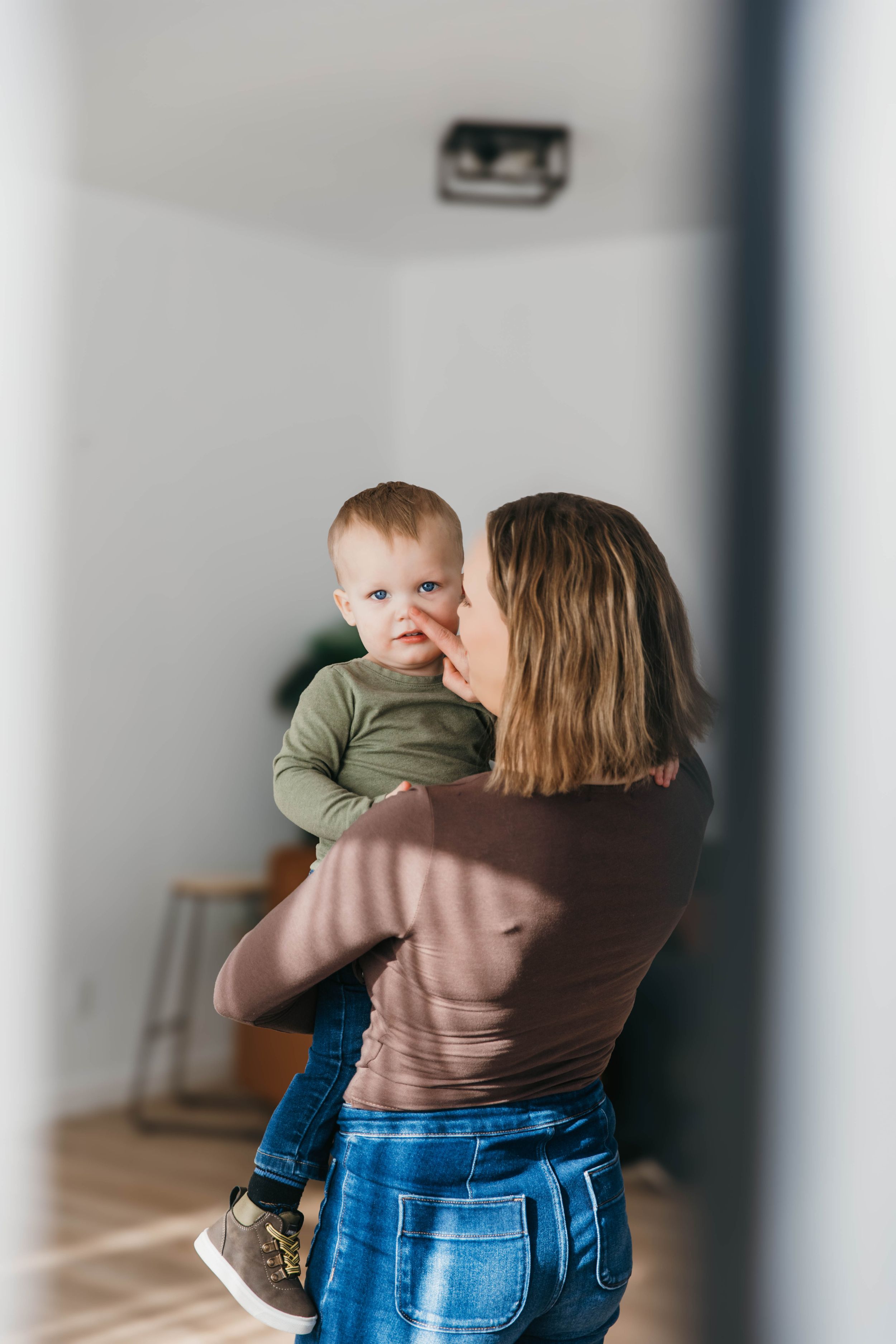 A parent in jeans and a brown sweater holds and spins with a young child wearing green in a bright indoor space.