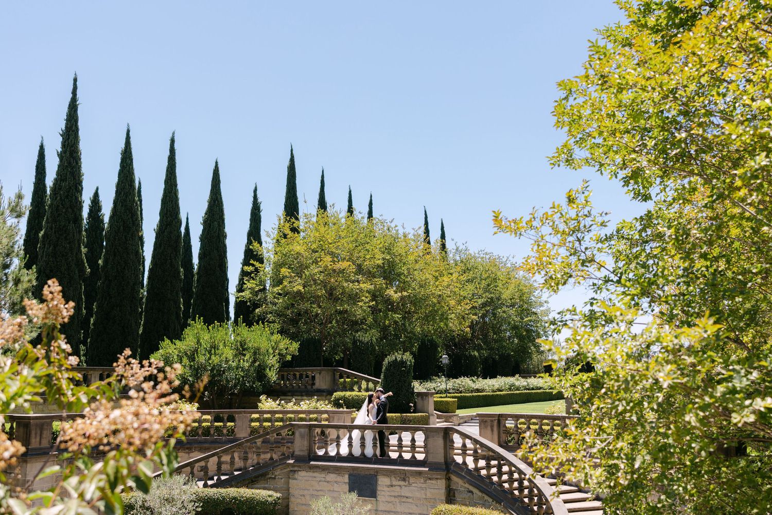 A couple stands on a stone balcony overlooking a lush garden with cypress trees and green landscaping on a sunny day.