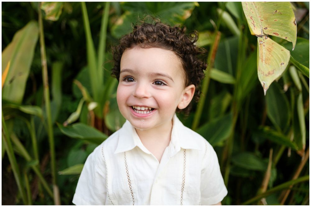 Young child in white shirt smiles brightly against lush green foliage background in outdoor portrait.