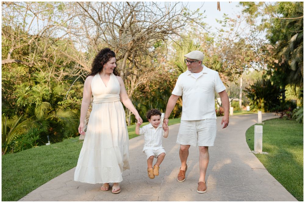 A family wearing white outfits walks together while holding hands on a paved garden path during a sunny day.