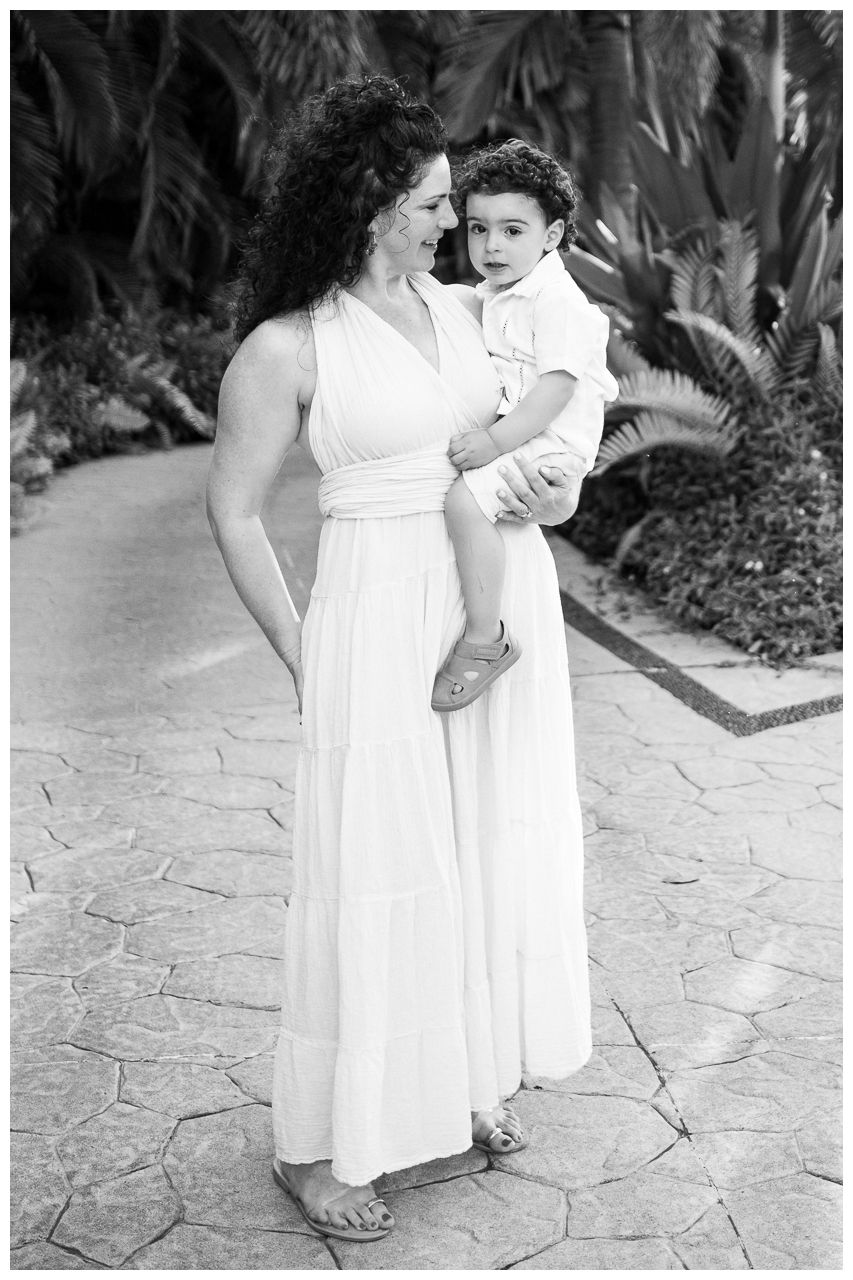 Black and white photograph of a parent in a flowing white dress holding a young child on a stone path with foliage behind them.