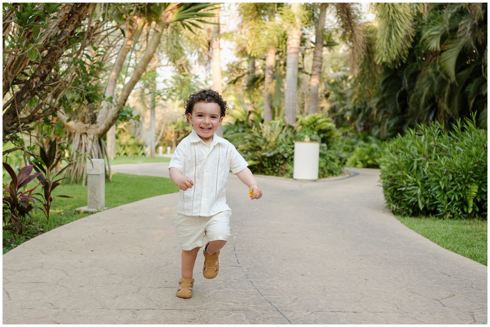 A young child in all-white outfit and tan boots runs joyfully down a palm-lined pathway.