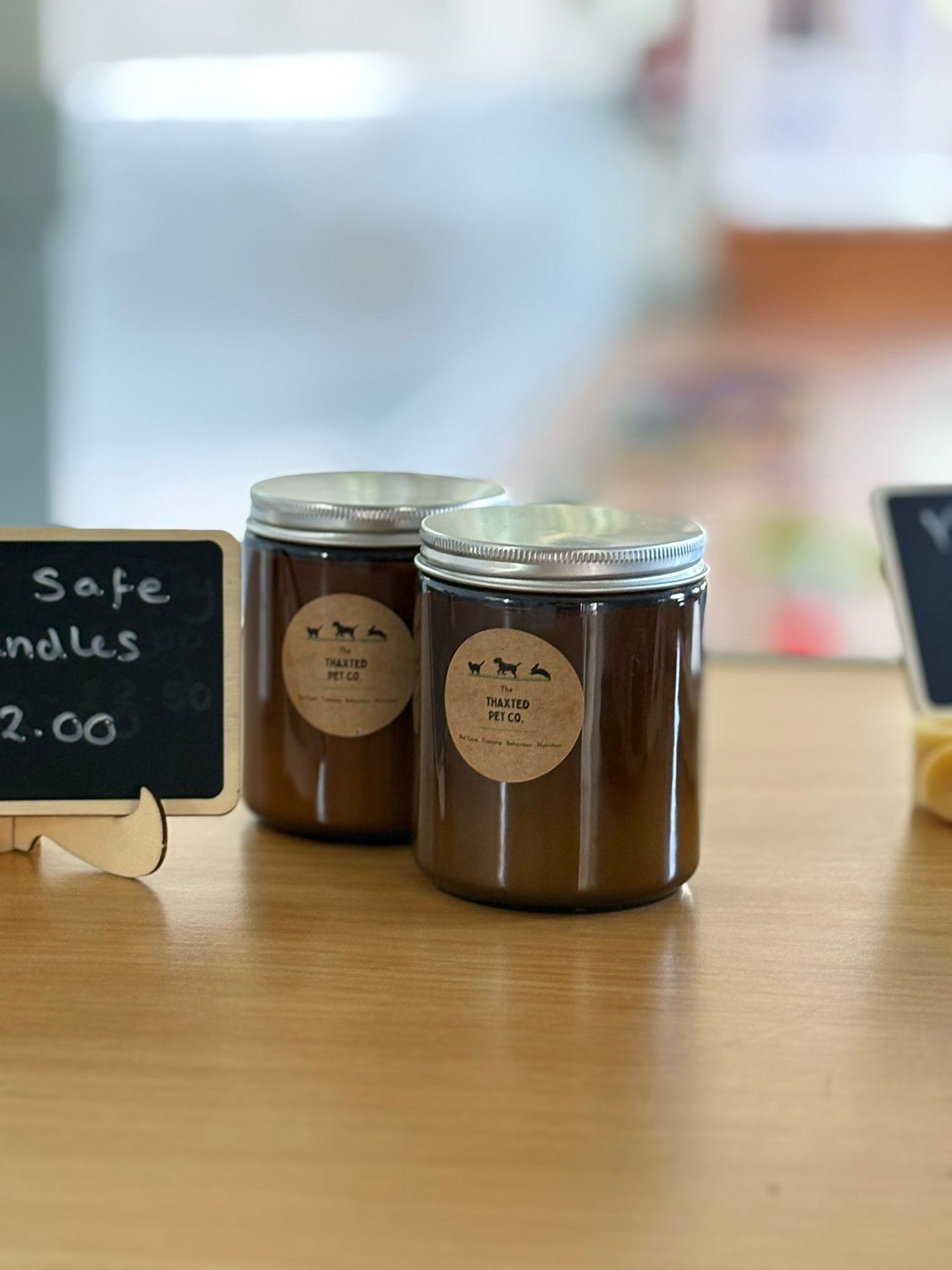 Two brown glass jar candles with rustic kraft paper labels sit on a wooden surface beside a small chalkboard price tag.