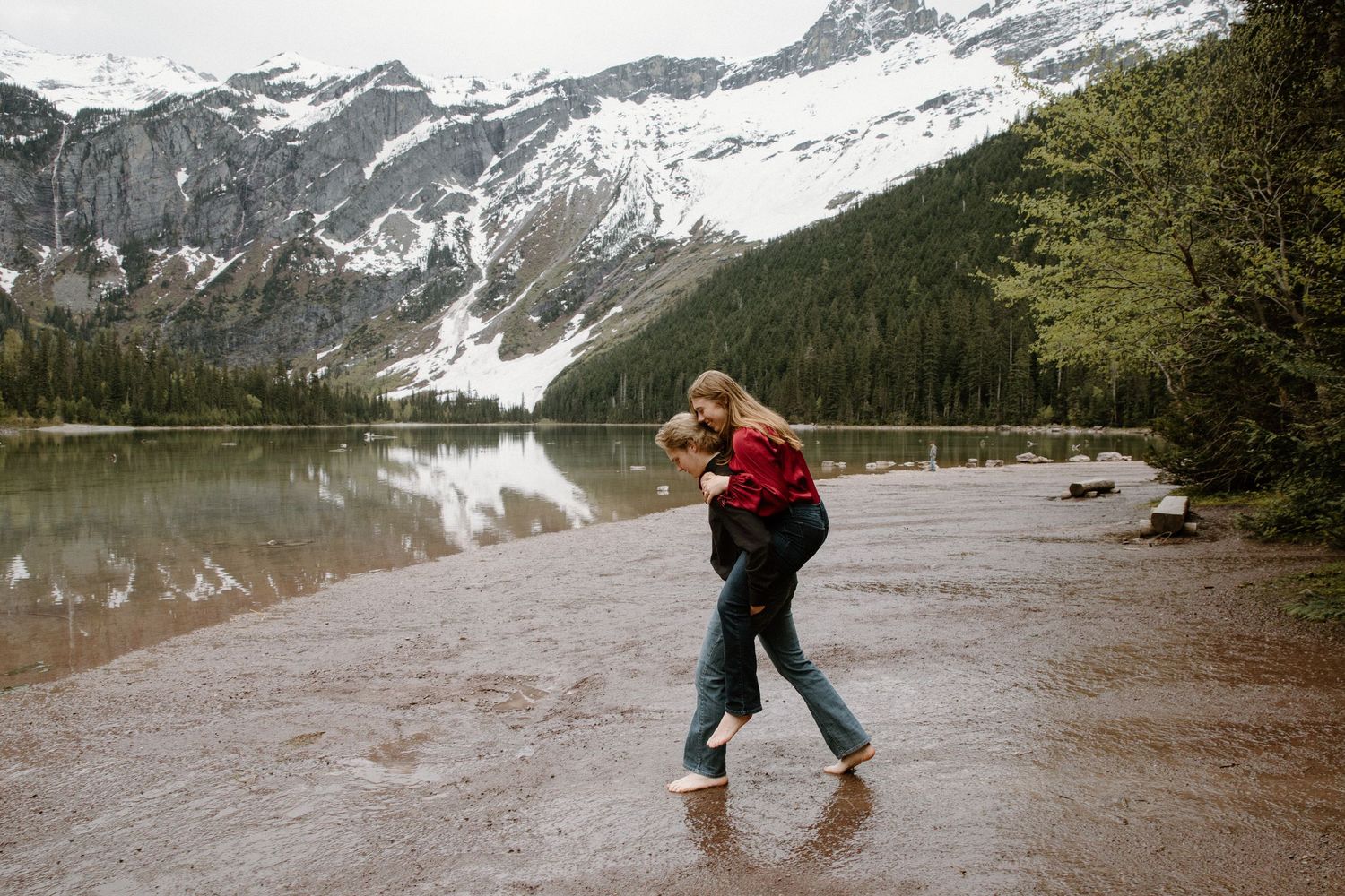 A person in a red sweater walks along a peaceful mountain lake shore with snow-capped peaks in the background.