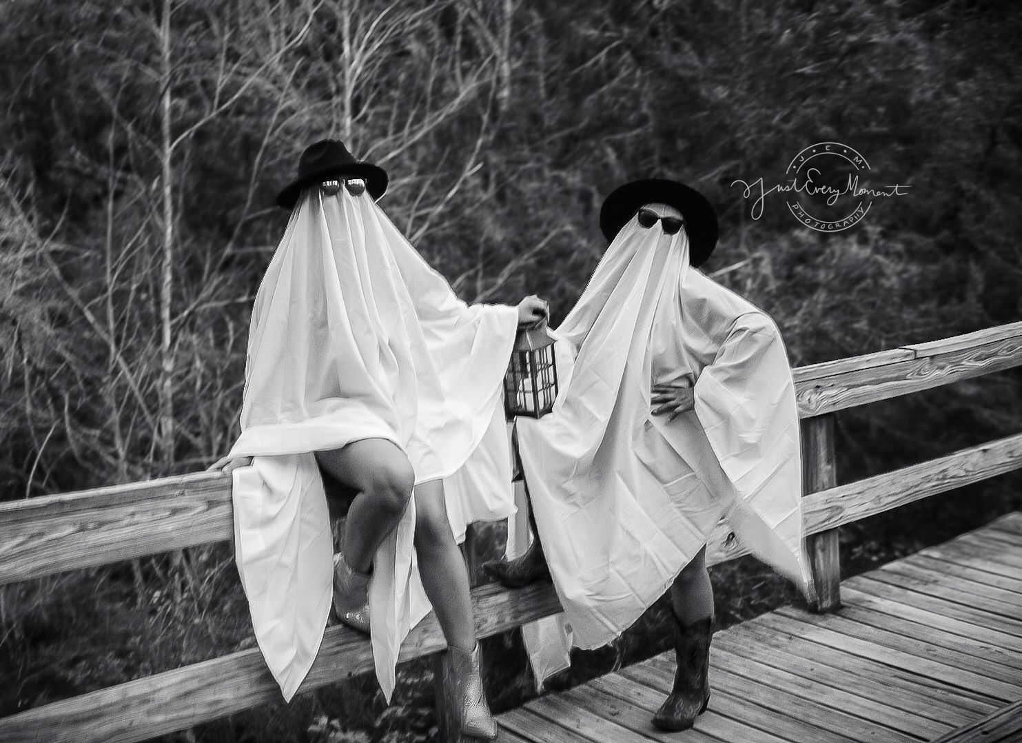 Two figures dressed as spooky ghosts with black hats lean against a wooden bridge railing in black and white.