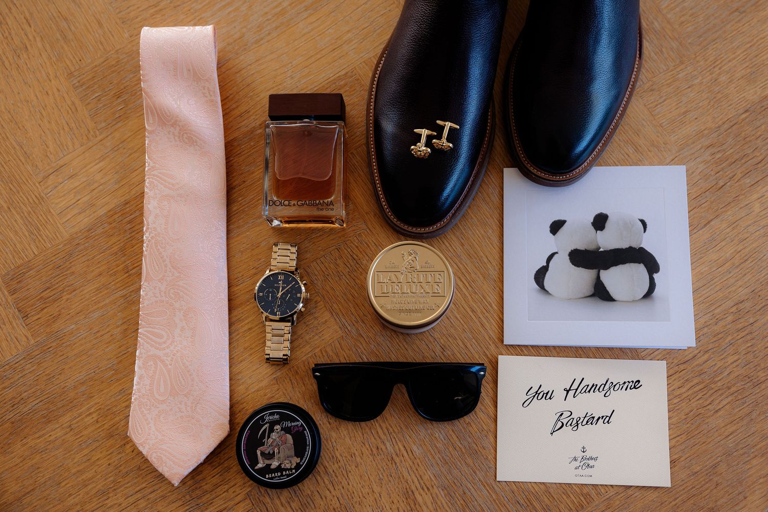 Groom's wedding accessories including peach tie, black dress shoes, sunglasses, watch, and cologne on wooden surface.