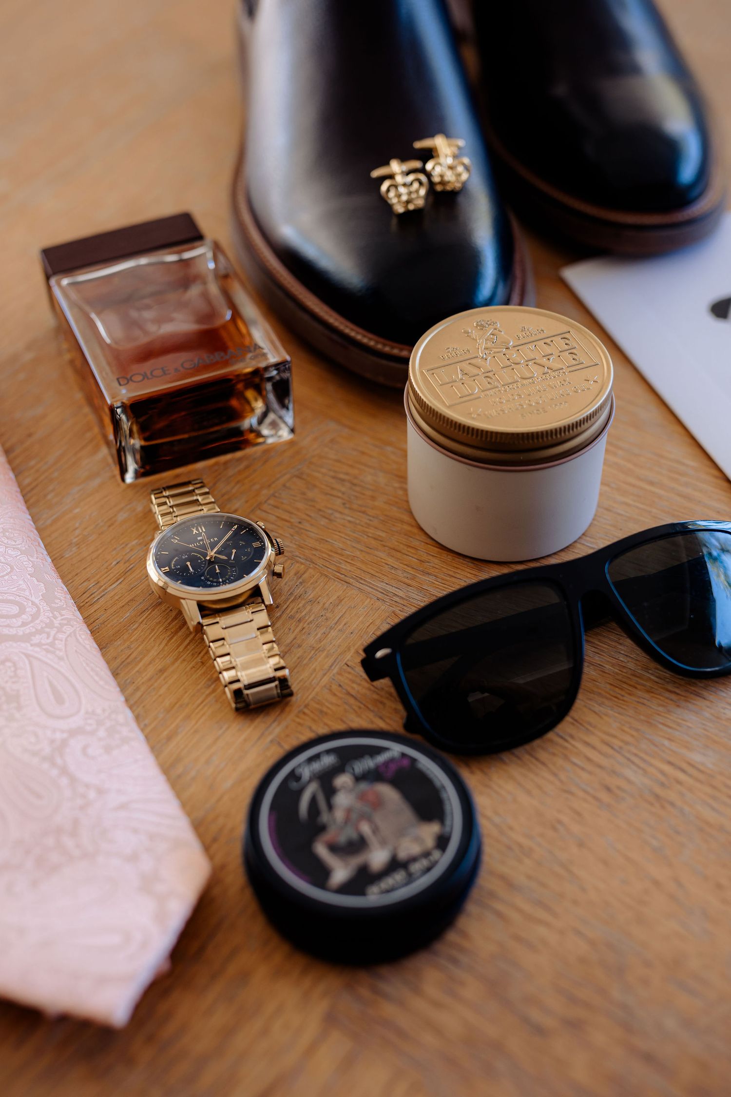 Men's fashion accessories including dress shoes, perfume, gold watch, sunglasses and grooming products on wooden surface.