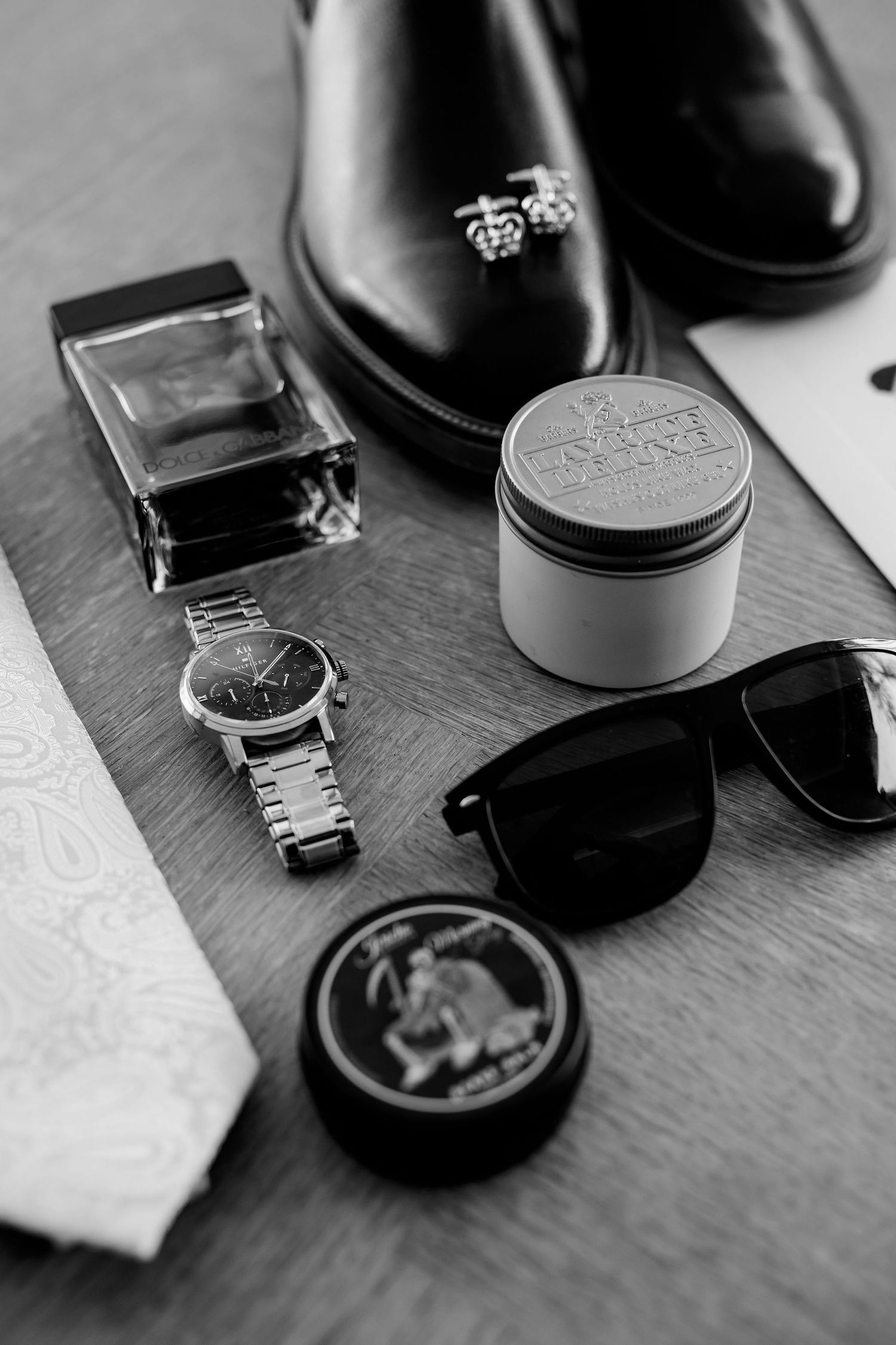 High-end men's accessories including dress shoes, sunglasses, watch, cologne and grooming products arranged on wooden surface.
