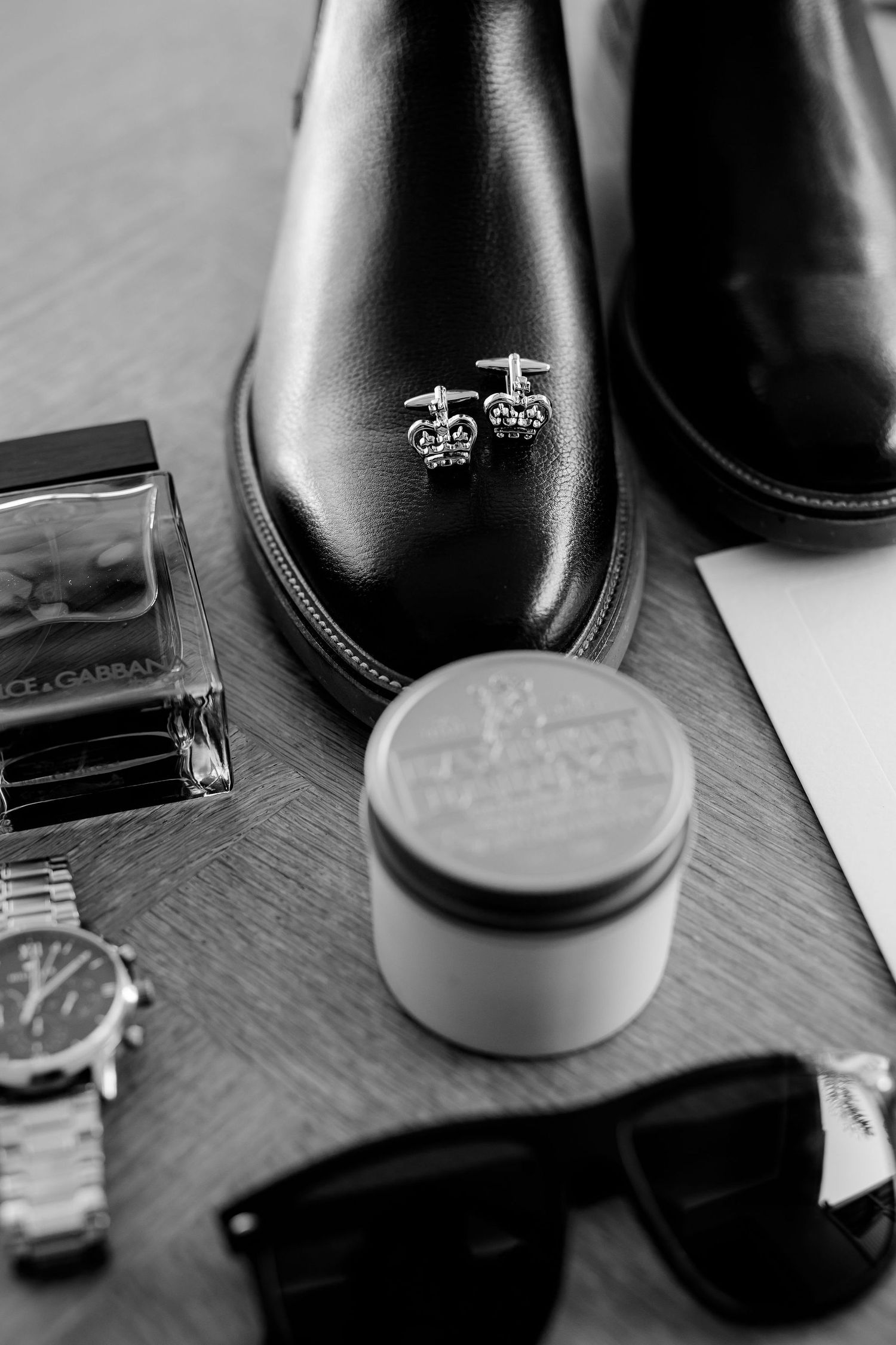 Black leather dress shoes with embroidered crown logo sit beside sunglasses, watch and grooming products on wooden surface.
