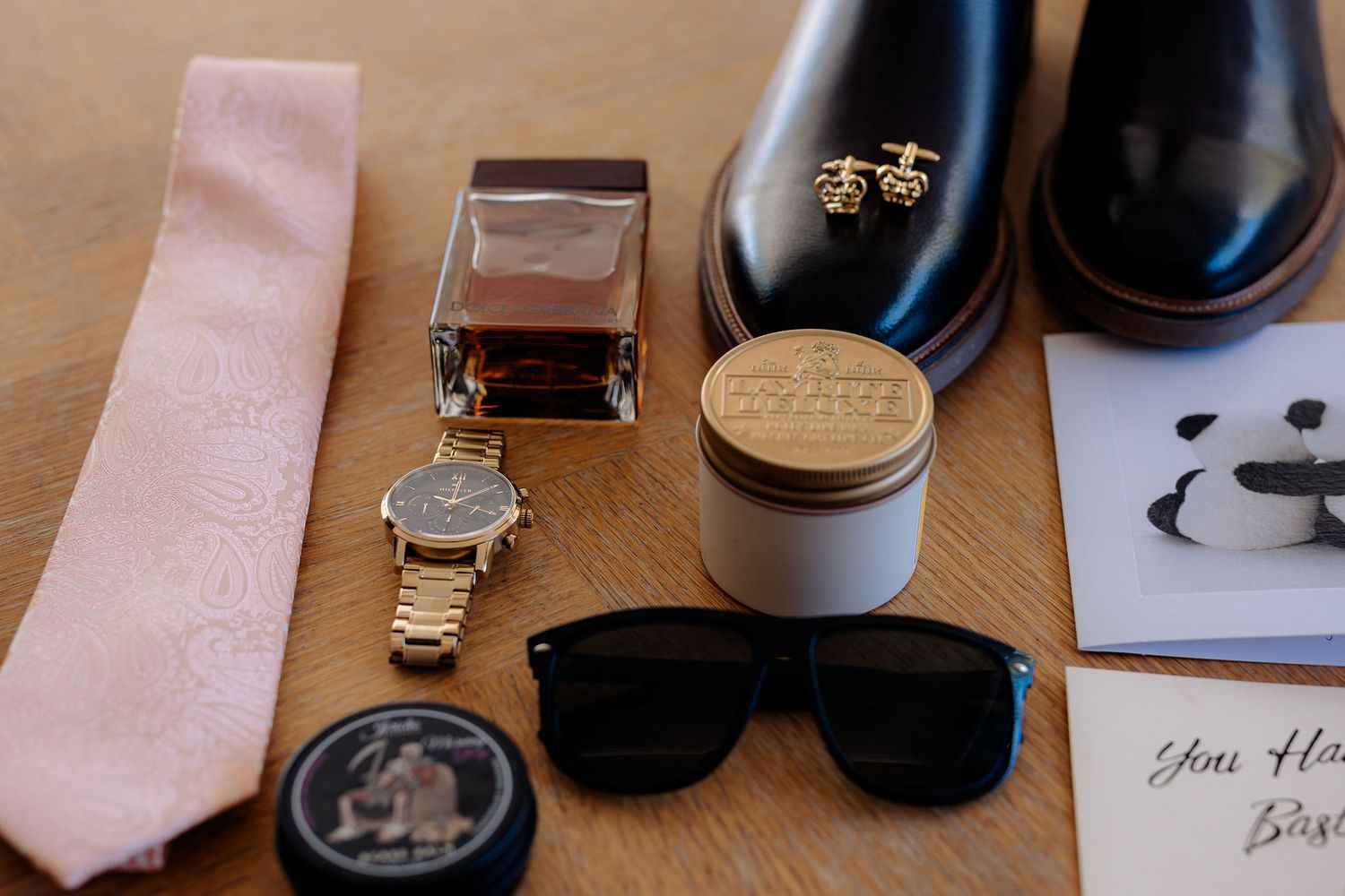 Wedding accessories including pink tie, watch, dress shoes, sunglasses and perfume laid out on brown surface.
