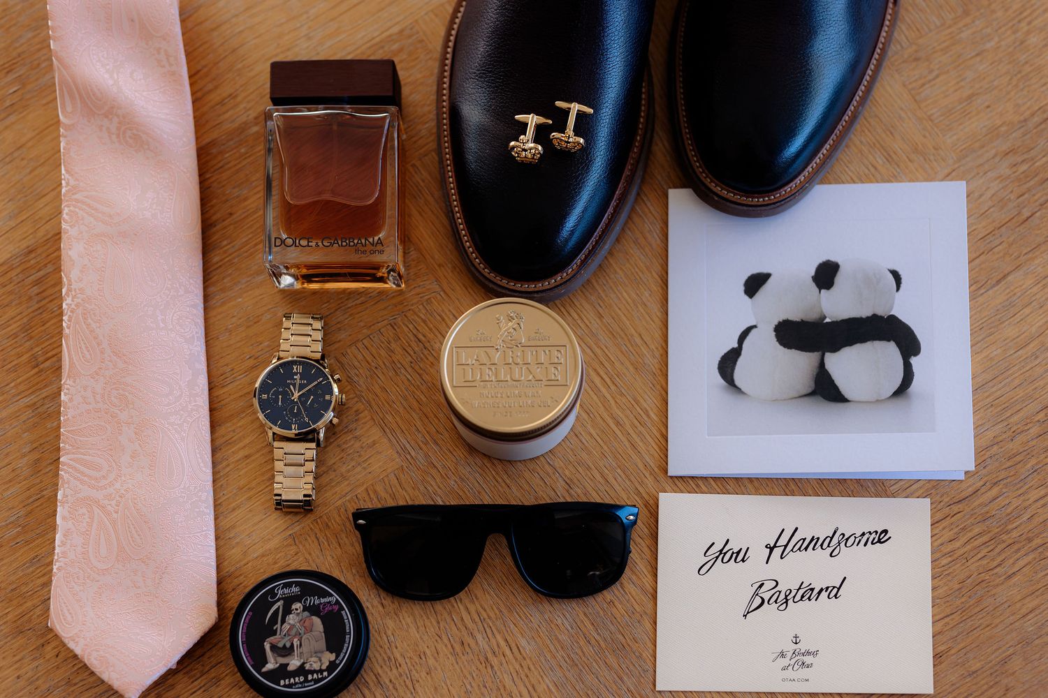 A flat lay of groom's accessories including sunglasses, shoes, perfume, cufflinks, watch, and pink tie on wooden surface.