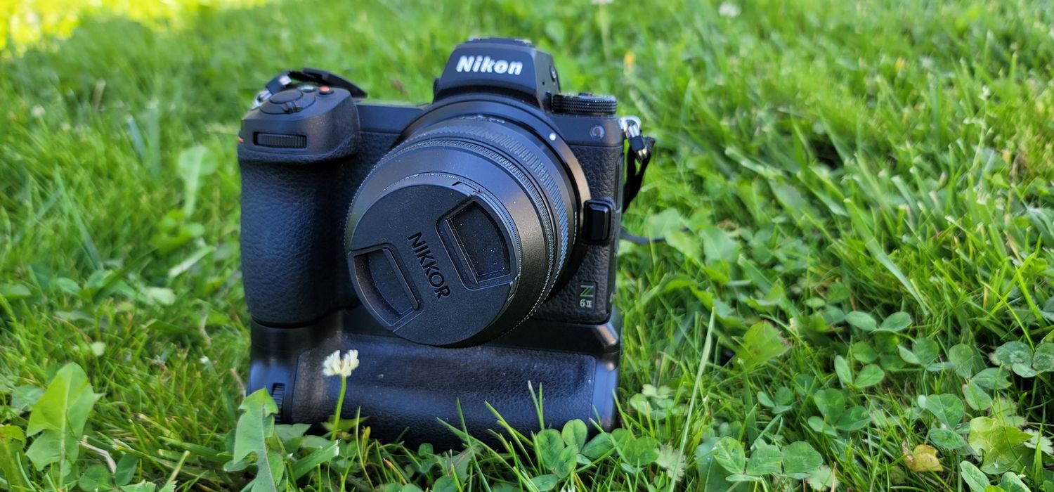 Nikon mirrorless camera with lens mounted on battery grip sits in green grass and clover.